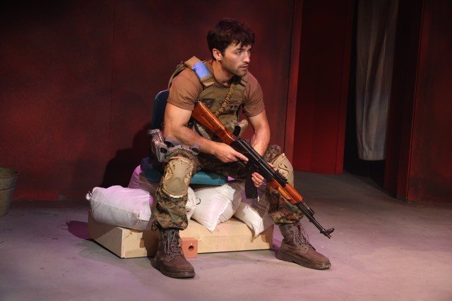 A soldier seated on sandbags, rifle across his lap, looking off stage