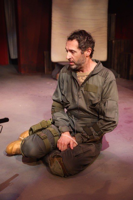 A soldier in a green jacket kneeling on the ground, mid-sentence